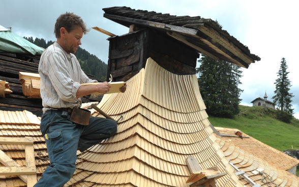 Ein Dachdecker erneuert den Schindelschirm eines traditionellen Alpgebäudes.